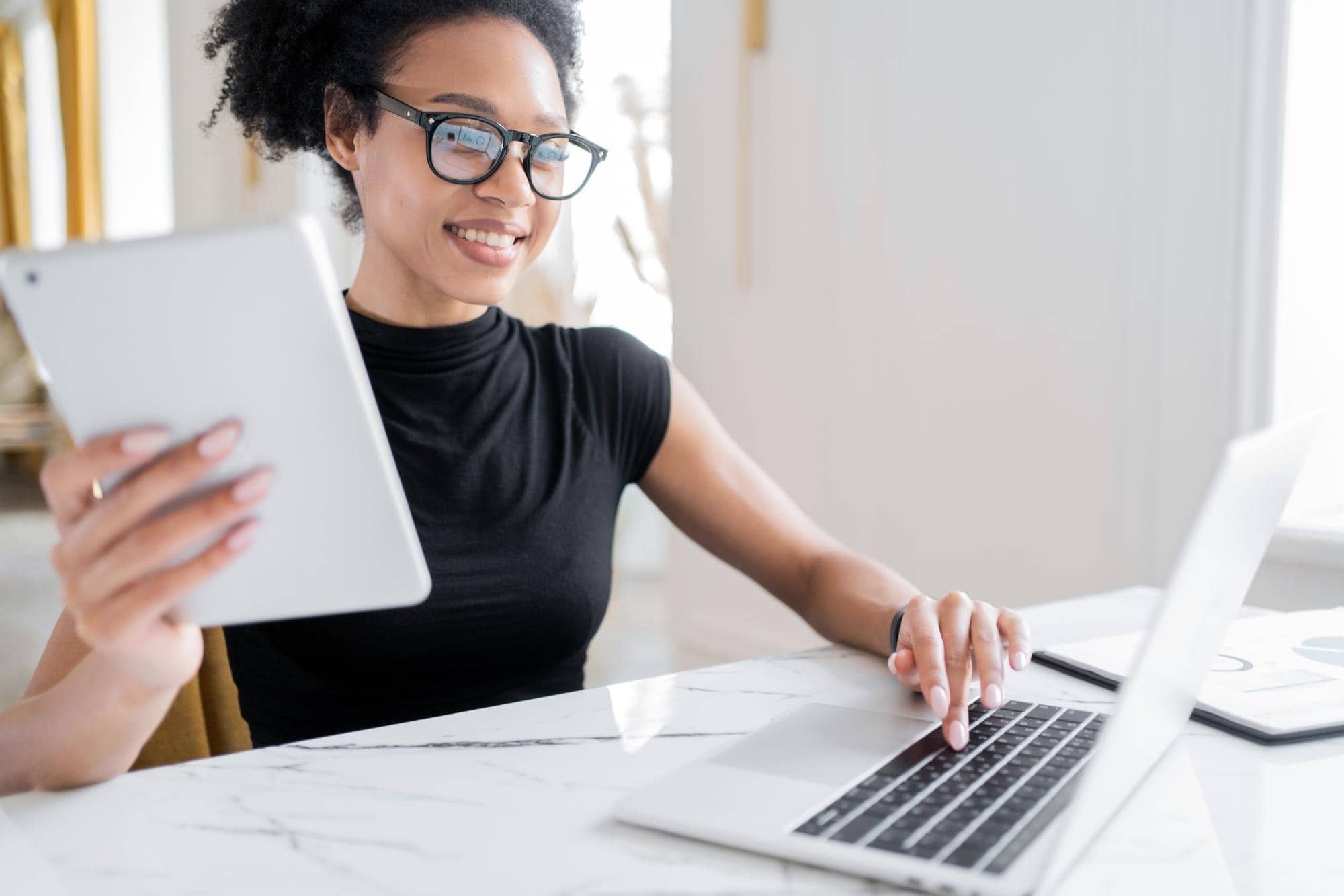 Bookkeeper reviewing monthly figures on a tablet and laptop, with a printed report on the desk