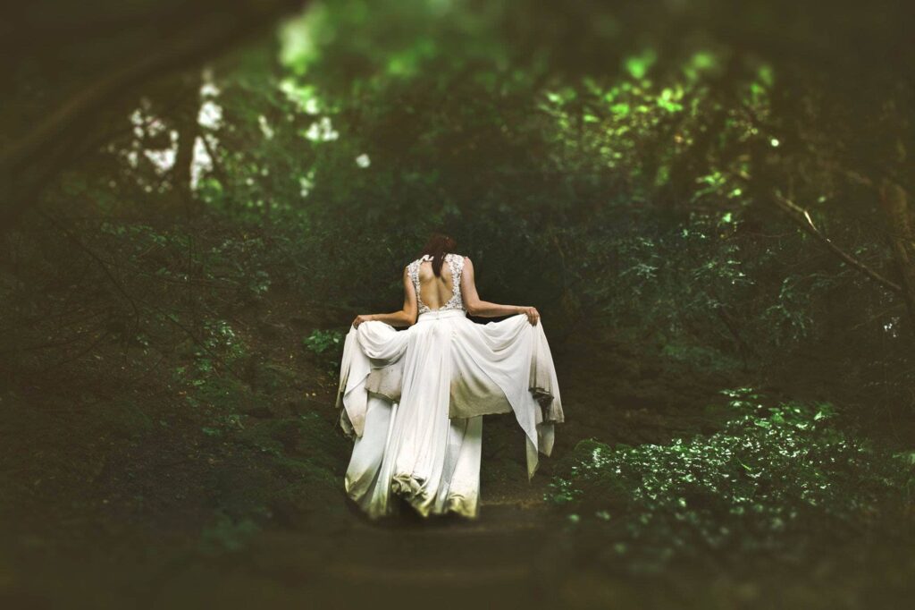 Woman in a long white gown walking into a dark green forest