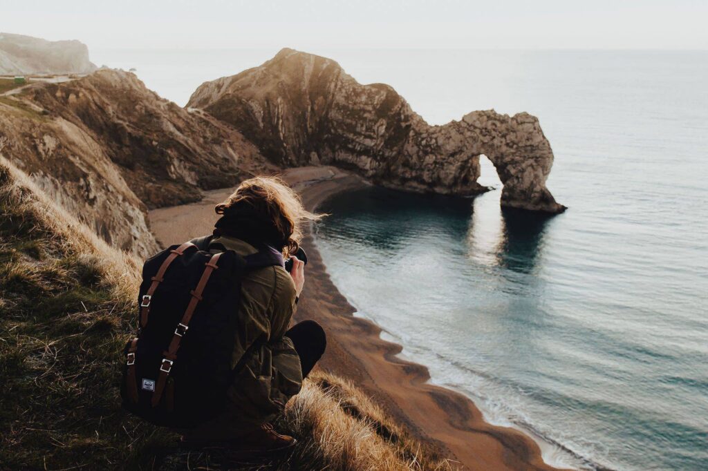 Photographer with a backpack on a clifftop above a sea arch at sunrise